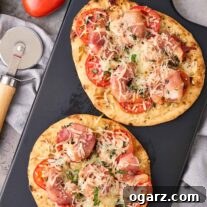 Two naan pizzas topped with prosciutto-wrapped shrimp, tomatoes, basil, and mozzarella on a black board with a pizza cutter.