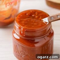 A small jar of bourbon barbecue sauce with a wooden-handled spoon in it, measuring cup and bottle of bourbon in the background.