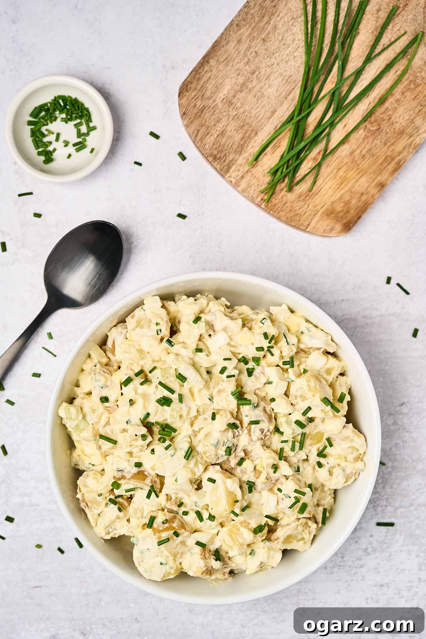 Sour Cream Potato Salad: A Zesty Twist on a Classic Side 2 Sour cream potato salad in a white serving bowl, garnished with fresh chives, a black serving spoon to the left, a prep bowl of chopped chives, and fresh chives on a wooden board in the background.