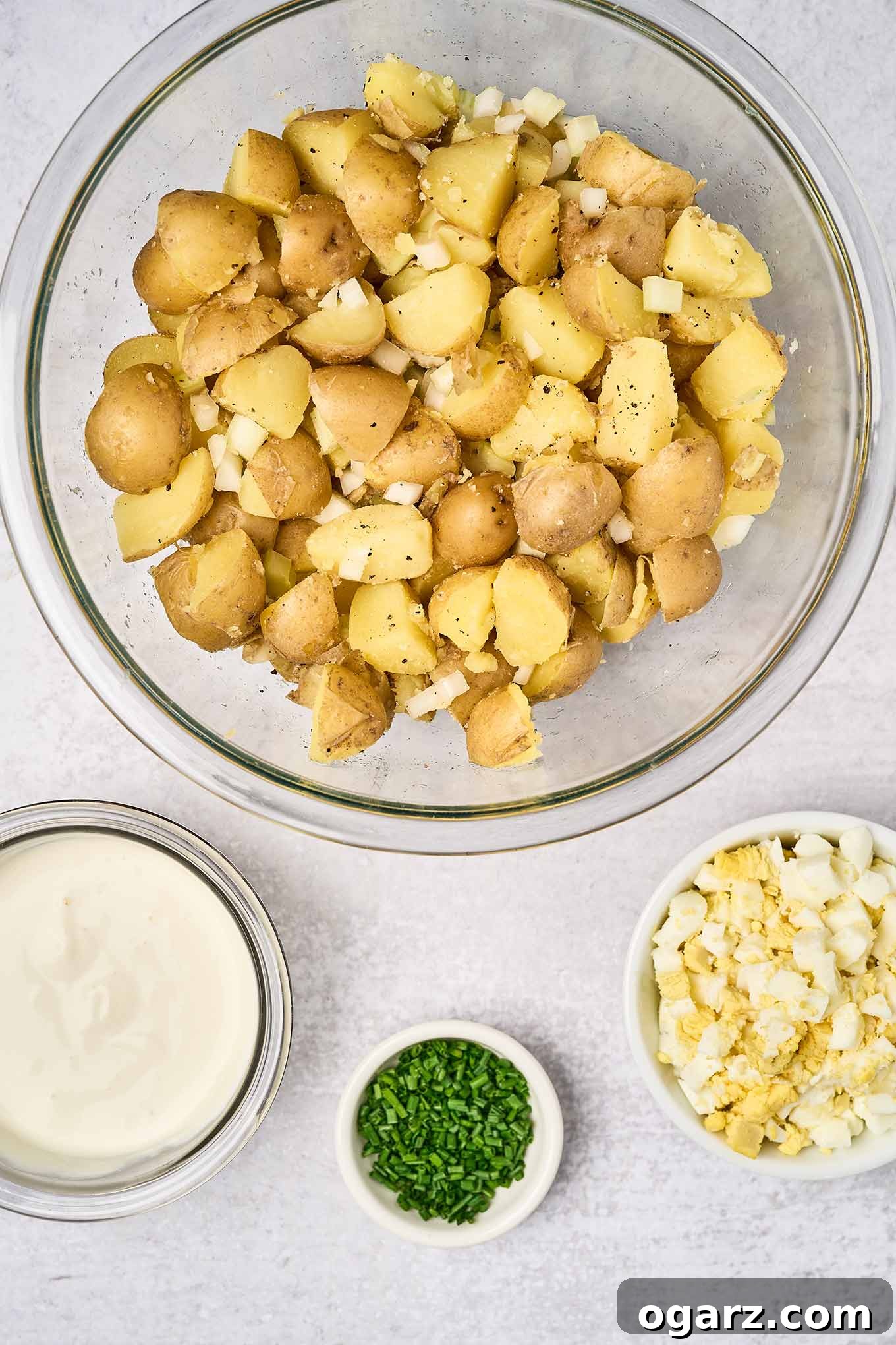 Sour Cream Potato Salad: A Zesty Twist on a Classic Side 4 Boiled chunks of baby yellow potatoes and chopped celery and onion in a mixing bowl next to a small bowl of chopped hard boiled eggs, a prep bowl of chives, and a bowl of sour cream dressing - all ready to be combined.