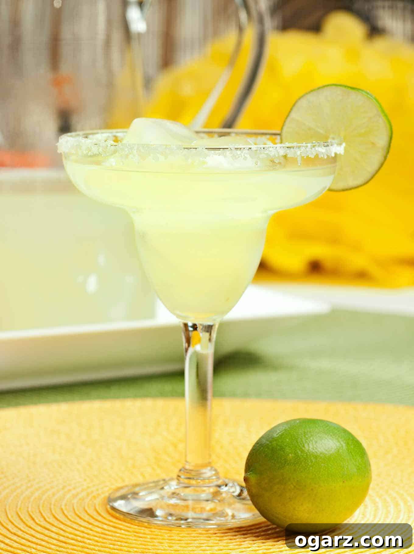 A perfectly balanced classic margarita on the rocks, garnished with a fresh lime wedge. A whole lime sits in the foreground, with a pitcher of more margaritas blurred in the background, ready for serving.