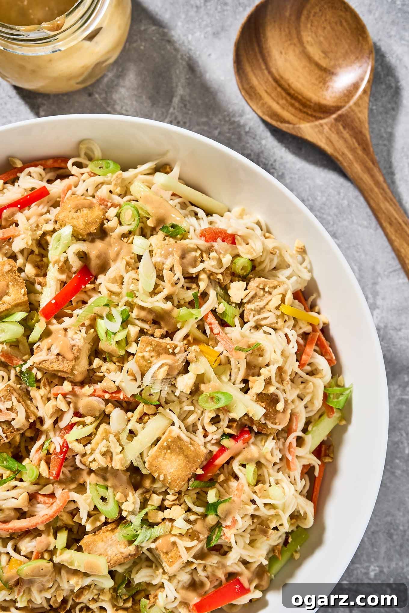 A vibrant serving bowl of Asian noodle salad featuring glistening ramen noodles, crispy golden tofu cubes, a medley of colorful shredded vegetables, and a rich peanut dressing, with extra dressing and salad servers placed to the side.