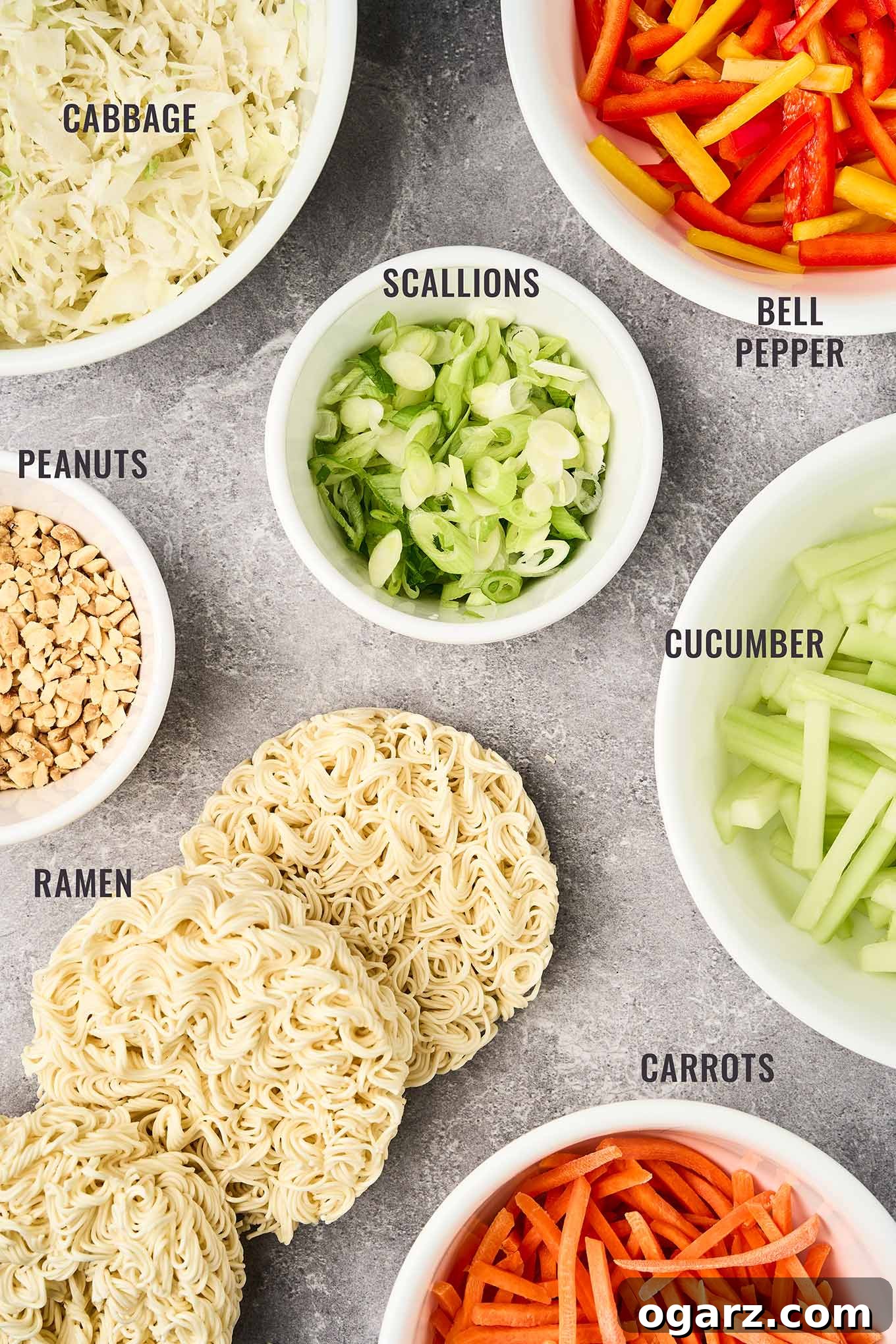 A labeled, overhead view of the vibrant ingredients needed to create a delicious Asian noodle salad, including shredded cabbage, colorful bell peppers, fresh scallions, crisp cucumber, cooked ramen noodles, roasted peanuts, and finely shredded carrots.