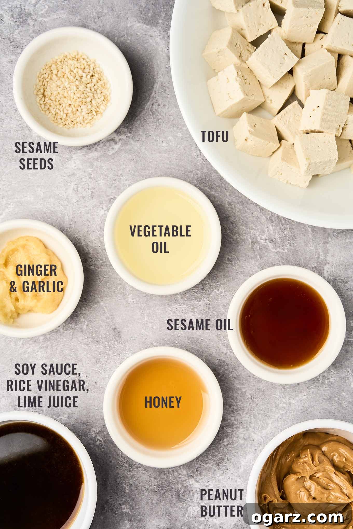 A labeled, overhead view of the essential ingredients for the creamy peanut dressing and crispy tofu in Asian noodle salad, featuring natural peanut butter, low-sodium soy sauce, golden honey, fresh lime juice, aromatic garlic, spicy ginger, firm tofu, and toasted sesame seeds.