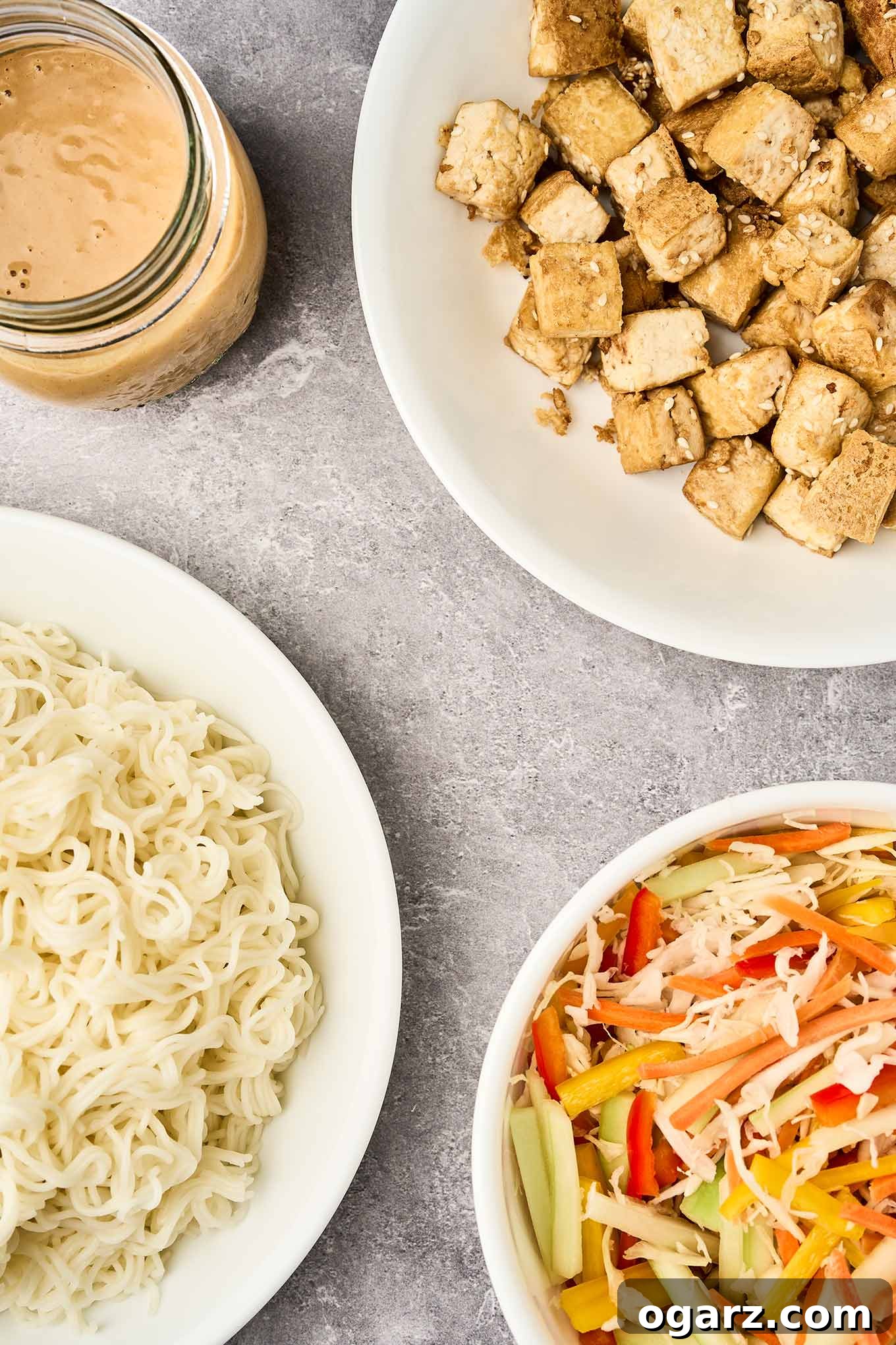All the components for the Asian noodle salad laid out and ready for assembly: a vibrant bowl of slivered bell peppers and cucumbers mixed with shredded carrots and cabbage, a separate bowl of perfectly cooked ramen noodles, a bowl of crispy golden tofu, and a jar of the homemade peanut dressing.