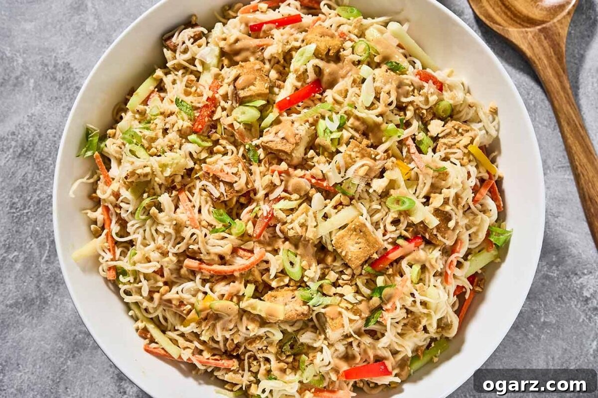 A vibrant and generously filled serving bowl of cold Asian noodle salad, perfectly tossed with a rich peanut dressing. The salad showcases a harmonious blend of ramen noodles, golden crispy tofu, shredded cabbage, colorful bell pepper strips, thin cucumber slices, grated carrots, fresh scallions, and a garnish of crushed peanuts and sesame seeds.