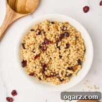 A bowl of toasted pearl couscous with dried cranberries and slivered almonds surrounded by a wooden spoon, scattered cranberries, and a wooden measuring cup with uncooked Israeli couscous.