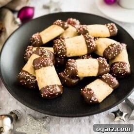 A black plate showcases delicious maple hazelnut cookies, with both ends elegantly dipped in chocolate and adorned with festive sprinkles, placed on a grey tablecloth. Nearby, silver and pink Christmas ornaments add to the holiday ambiance.