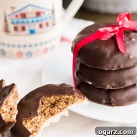 A stack of three chocolate-covered Lebkuchen with a red bow on a white plate, next to a Lebkuchen broken in half. A cozy cup is visible in the background.
