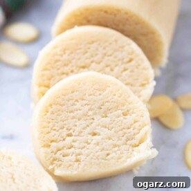 A smooth log of homemade marzipan, with two perfectly cut slices leaning against it, displayed on a grey marble surface alongside a scattering of raw almonds.