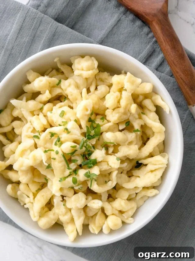 Effortless Homemade German Spaetzle 2 Top-down shot of a white bowl of spaetzle garnished with parsley on a grey dishtowel next to a wooden spoon.