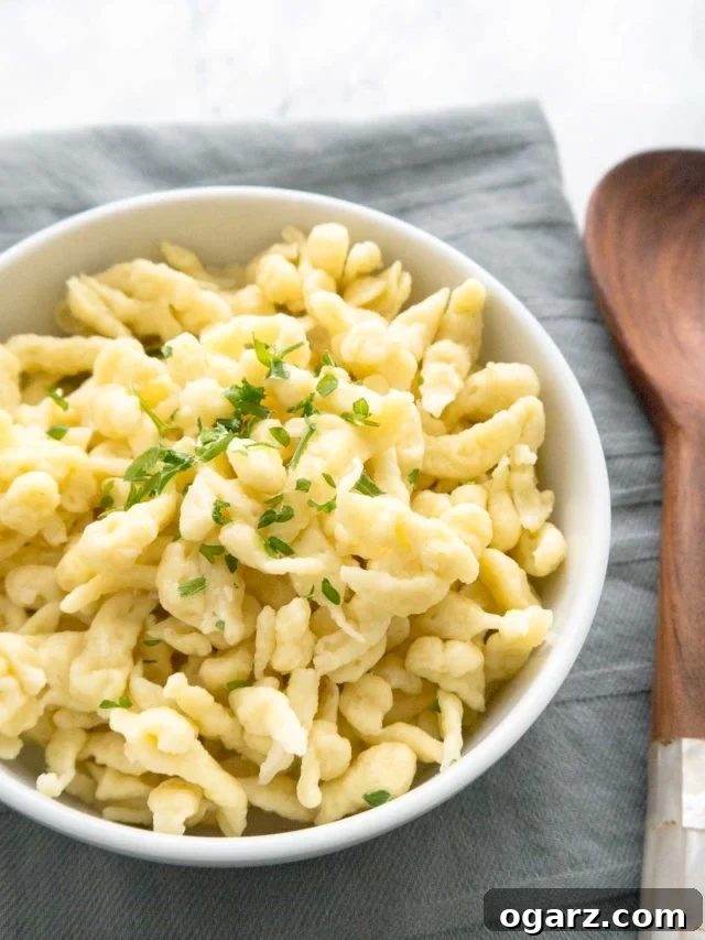 Effortless Homemade German Spaetzle 3 A white bowl of spaetzle garnished with parsley on a grey dishtowel next to a wooden spoon.