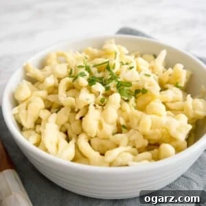 Effortless Homemade German Spaetzle 6 A white bowl of spaetzle garnished with parsley on a grey dishtowel next to a wooden spoon.