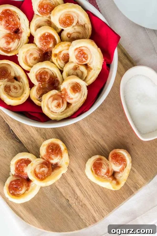 These heart-shaped Palmiers are made with only two ingredients! A French cookie recipe that's special enough for Valentine's Day but easy enough for every day. These puff pastry cookies are loved by kids and adults alike! #valentinesdaytreats #cookies #puffpastry #valentinesdesserts Top-down shot of heart-shaped puff pastry palmiers. Three are on a cutting board and the rest are in a white bowl lined with a red napkin.