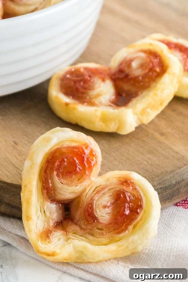 These heart-shaped Palmiers are made with only two ingredients! A French cookie recipe that's special enough for Valentine's Day but easy enough for every day. These puff pastry cookies are loved by kids and adults alike! #valentinesdaytreats #cookies #puffpastry #valentinesdesserts Close-up of a heart-shaped puff pastry palmier leaning against a wood cutting board with more palmiers in the background.