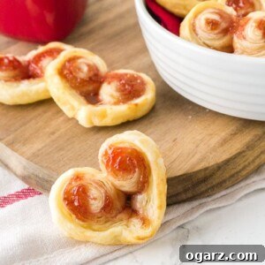 Heart-shaped puff pastry palmiers. One is in the front leaning against a wooden cutting board, others are lying on the cutting board and in a white bowl on the board.