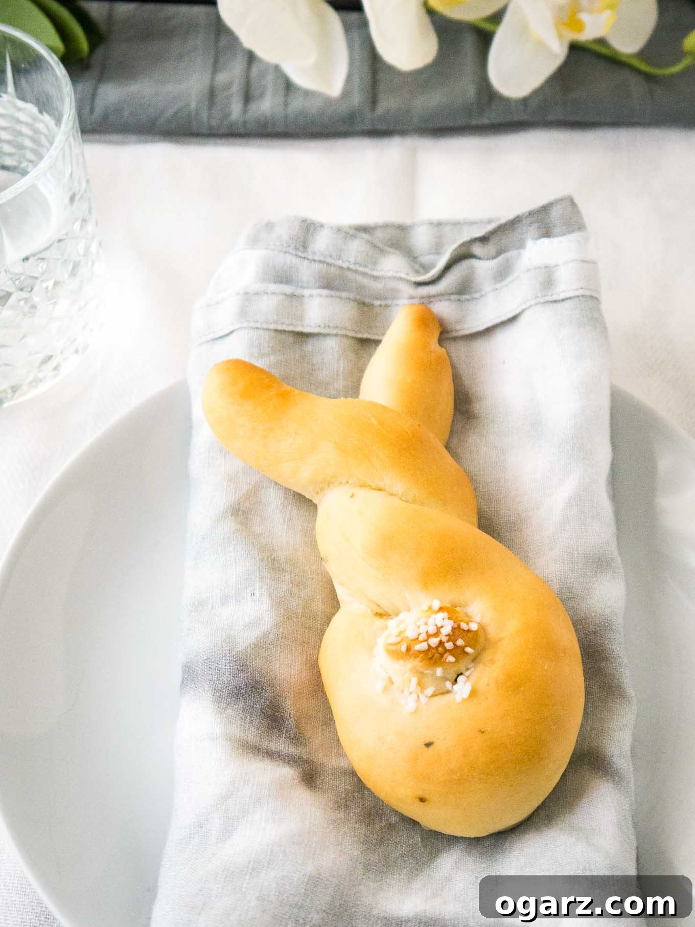 Sweet Easter Bunny Delights 2 Golden-brown bunny-shaped bread rolls with small dough cottontails, arranged on a grey dishtowel next to a glass of water on a white plate, ready to be served.