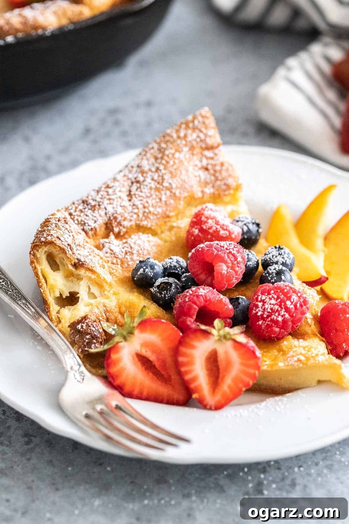Spectacular Oven-Baked Pancake 2 A slice of Dutch Baby pancake on a white plate garnished with fresh berries and a dusting of powdered sugar.