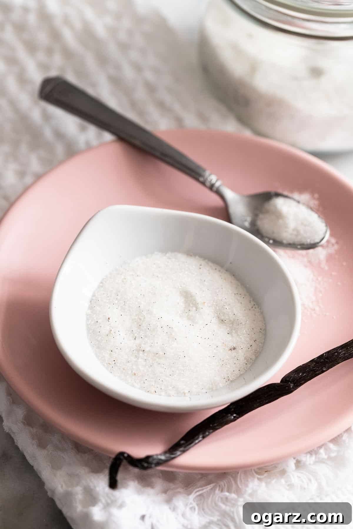 A small bowl filled with vanilla sugar on a pink plate with a spoon and vanilla bean next to it.