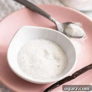 A small bowl filled with vanilla sugar on a pink plate with a spoon and vanilla bean next to it.