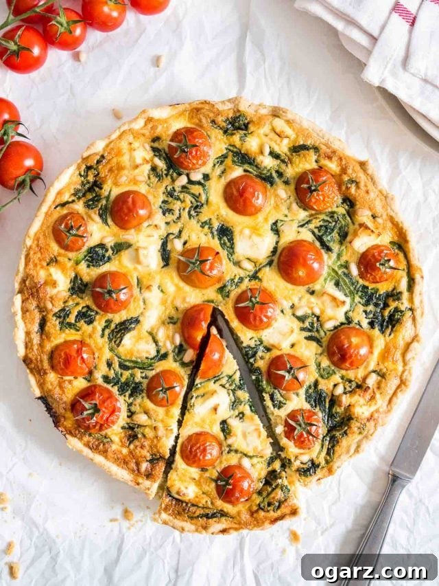 Spinach quiche with tomatoes sitting on a white dishtowel with a knife, a red and white dishtowel and some tomatoes next to it. A slice has been cut but is still in place.