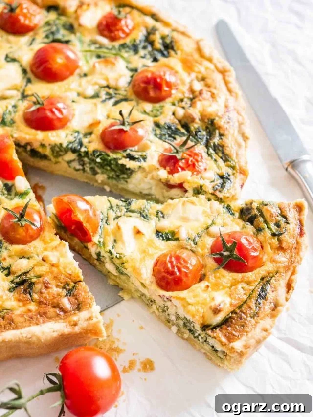 Close-up of spinach quiche with tomatoes sitting on a white dishtowel with a knife and some tomatoes next to it. A slice has been cut and pulled out slightly.