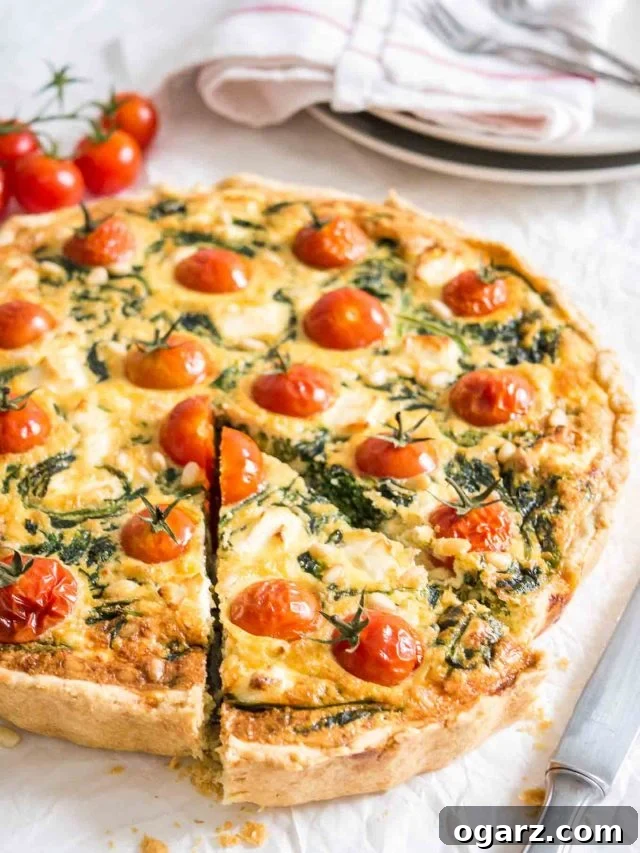 Close-up of spinach quiche with tomatoes sitting on a white dishtowel with a knife, a red and white dishtowel and some tomatoes next to it. A slice has been cut but is still in place.