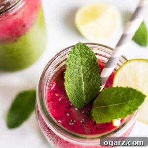 A jar of coconut water smoothie, with raspberries, baby spinach, chia, and mango, garnished with mint and a slice of lime with a straw in it. There's another glass of the smoothie an limes and mint next to it.