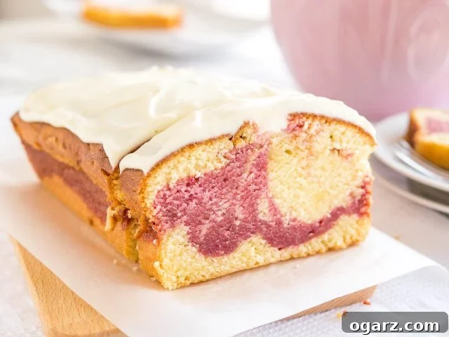 A delightful loaf of raspberry swirl pound cake, recently baked, rests elegantly on a wooden cutting board lined with parchment paper. A single slice has been carefully removed and is propped against the main loaf. In the background, there is a charming pink cup and a neat stack of two white plates, with another slice of the delicious cake ready for enjoyment.