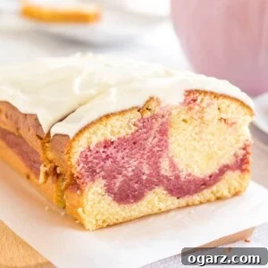 A loaf of raspberry swirl pound cake sitting on a wooden cutting board lined with parchment paper. A slice has been cut off and is leaning against it. In the background, there's a pink cup and a stack of two plates with another slice on it.