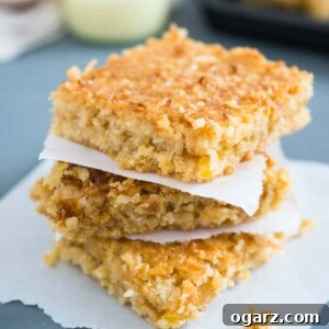 A stack of tropical coconut squares on a piece of parchment paper. There's a glass of sweetened condensed milk in the background next to a baking sheet with the rest of the squares.