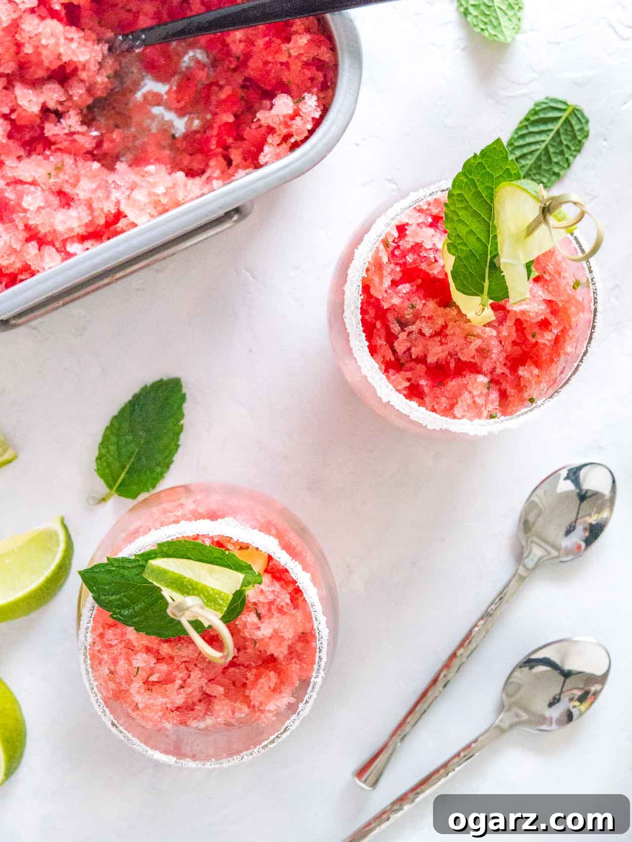 Slushy Watermelon Delight 4 Top-down shot of two sugar-rimmed glasses of pink watermelon granita with skewers with lime and mint. There are spoons, lime wedges, mint and a stainless steel baking pan of granita next to it.