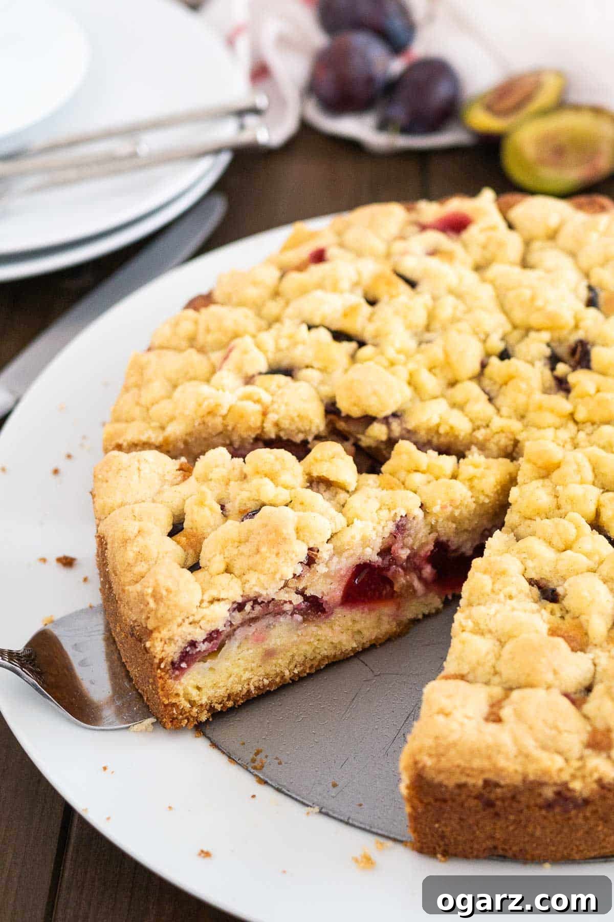 A plum cake with streusel on a springform bottom on a white plate. A cake server is lifting out a slice.