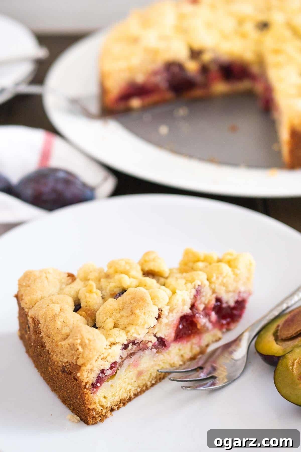 A slice of plum cake topped with streusel on a white plate with a fork and a halved plum. The whole cake is in the background.