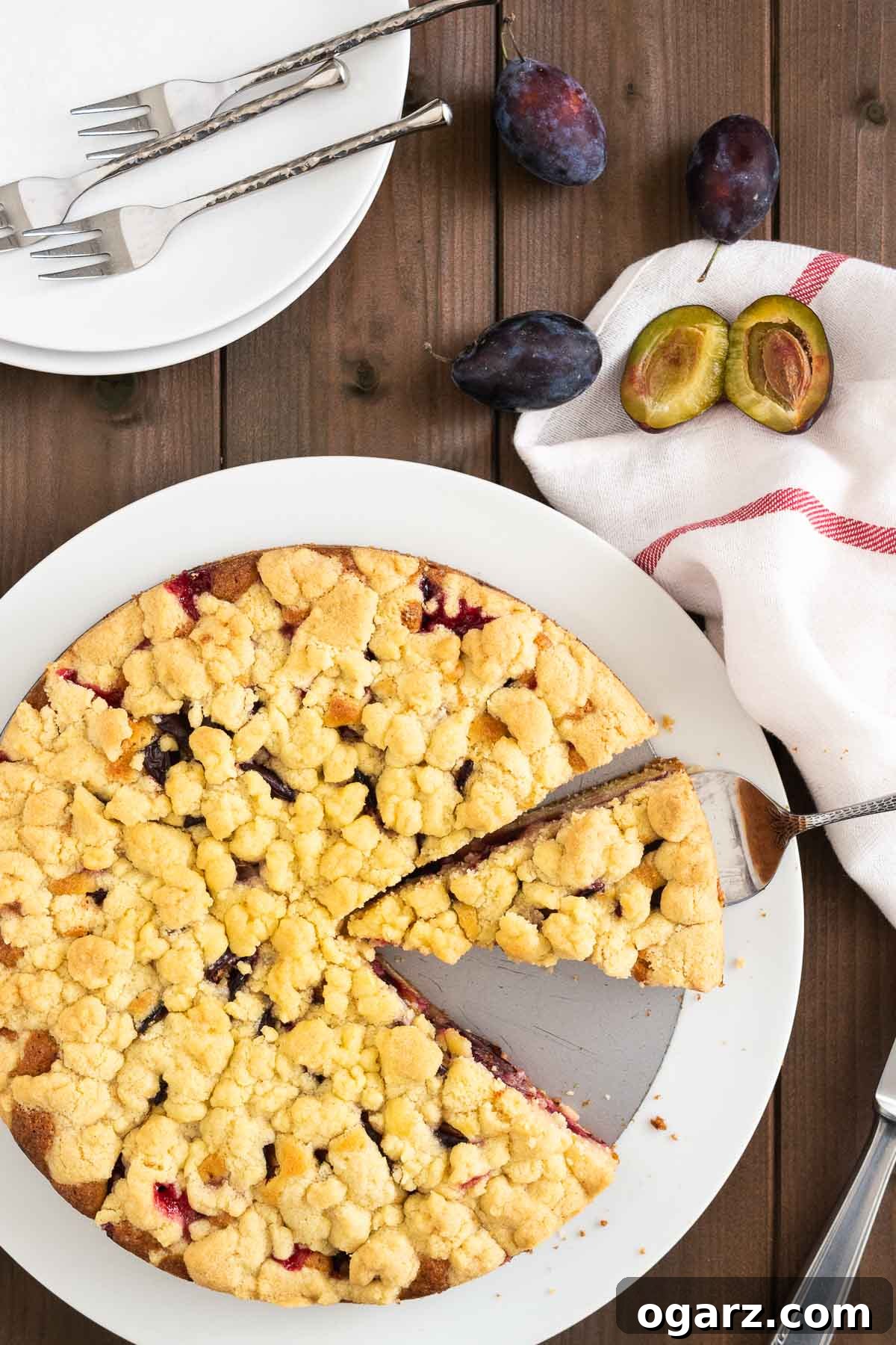 Top-down shot of a plum cake with streusel on a springform bottom on a white plate. A cake server is lifting out a slice. Next to the plate, there are plums, a knife, two white plates with forks and a white dishtowel with a red stripe.