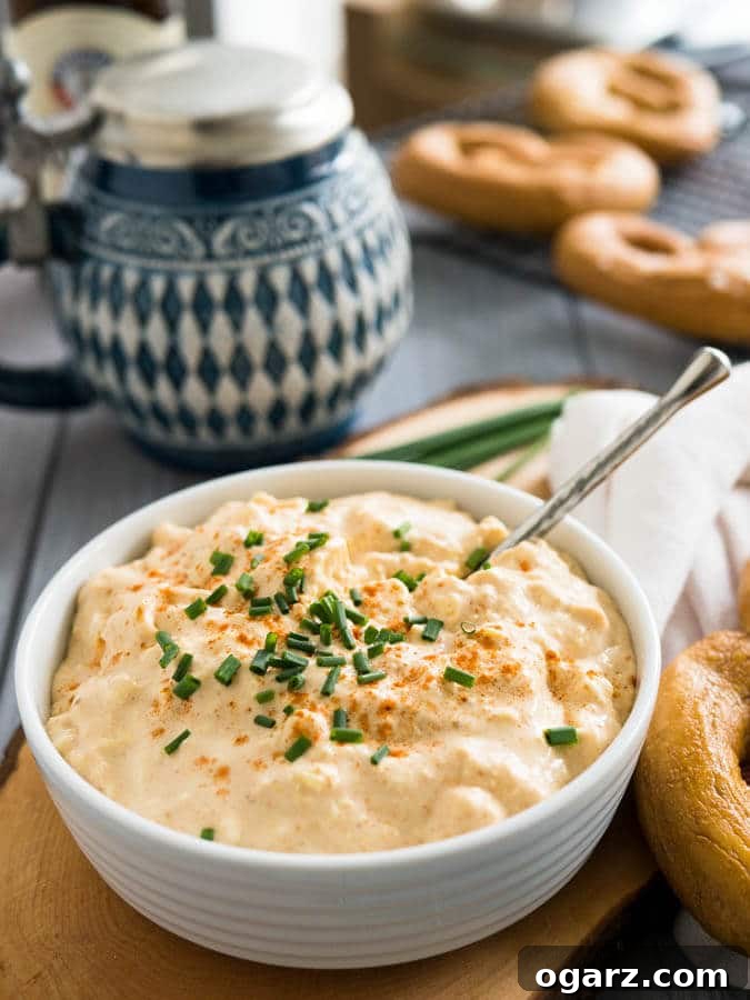 Obatzda Creamy Bavarian Beer Cheese Spread 4 A white bowl of Obatzda (German Beer Cheese Dip) with a spoon, garnished with chives on a wooden cutting board. There's a pretzel on a white dishtowel with a red stripe next to it and a beer stein in the background with more pretzels.