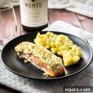 A black plate with parmesan-crusted salmon and mashed potatoes on a grey dishtowel on a wooden table. There's a glass and a bottle of wine next to it.