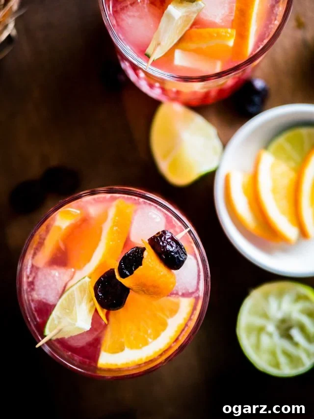 This Orange Cranberry Moscow Mule is the perfect holiday drink! A festive spin on a traditional cocktail made with cranberry juice, fresh oranges, and lime. Ahşap bir masa üzerinde, meyve şişleriyle süslenmiş bir Orange Cranberry Moscow Mule'un tepeden çekilmiş fotoğrafı. Yanında bir bardak daha aynı kokteyl ve içinde limon ve portakal dilimleri olan beyaz bir kase bulunuyor.