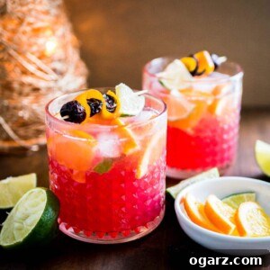 Crimson Citrus Moscow Mule 2 Parlak kırmızı ve turuncu renkli iki bardak Cranberry Orange Moscow Mule, portakal ve kızılcık dilimleri ile süslenmiş. Arka planda limon ve portakal dilimleri ile bir Noel süsü görülüyor.