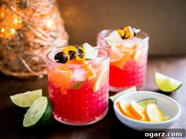 This Orange Cranberry Moscow Mule is the perfect holiday drink! A festive spin on a traditional cocktail made with cranberry juice, fresh oranges, and lime. Parlak kırmızı ve turuncu renkli iki bardak Cranberry Orange Moscow Mule, portakal ve kızılcık dilimleri ile süslenmiş. Arka planda limon ve portakal dilimleri ile bir Noel süsü görülüyor.