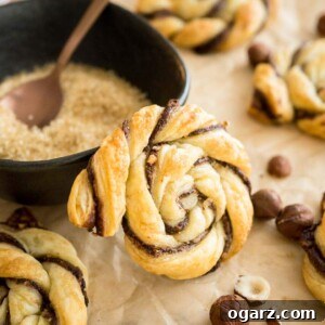 Simple Swirl Nutella Danish 8 A twisted nutella danish leaning against a black bowl with brown sugar and a bronze spoon garnished with hazelnuts on brown parchment paper. There are more of the danishes around it.