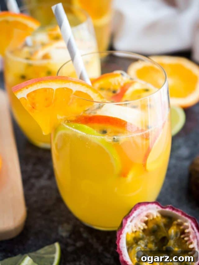 Close-up of a glass of tropical white wine sangria with orange slices, passion fruit seeds and a straw on a dark surface. There are orange slices, another glass and passion fruits next to it.