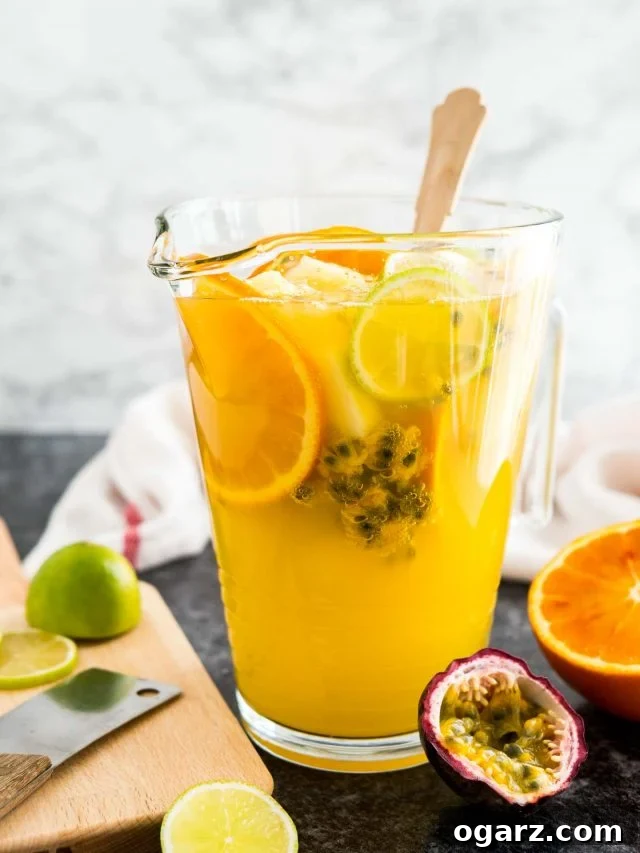 A glas pitcher or tropical white wine sangria with a wooden spoon, orange slices and passion fruit. There are limes, passion fruits and oranges next to it as well as a wooden cutting board and a white dishtowel.