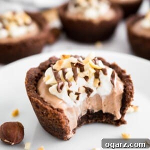 Decadent Nutella Mousse Cookie Cups 5 A white plate with a Nutella Mousse Cookie Cup, a bite taken out, garnished with hazelnuts. More cookie cups are in the background.