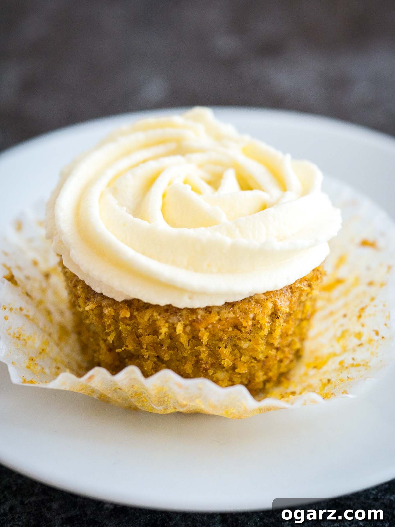 A perfectly frosted carrot cake cupcake with creamy white frosting, nestled in a decorative paper liner on a pristine white plate, ready to be enjoyed.