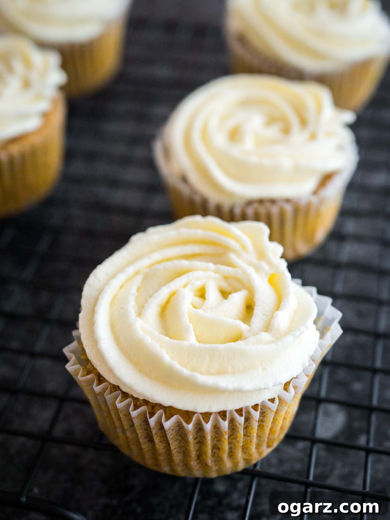 A striking display of carrot cake cupcakes, topped with elegant swirls of cream cheese frosting, resting on a dark cooling rack.