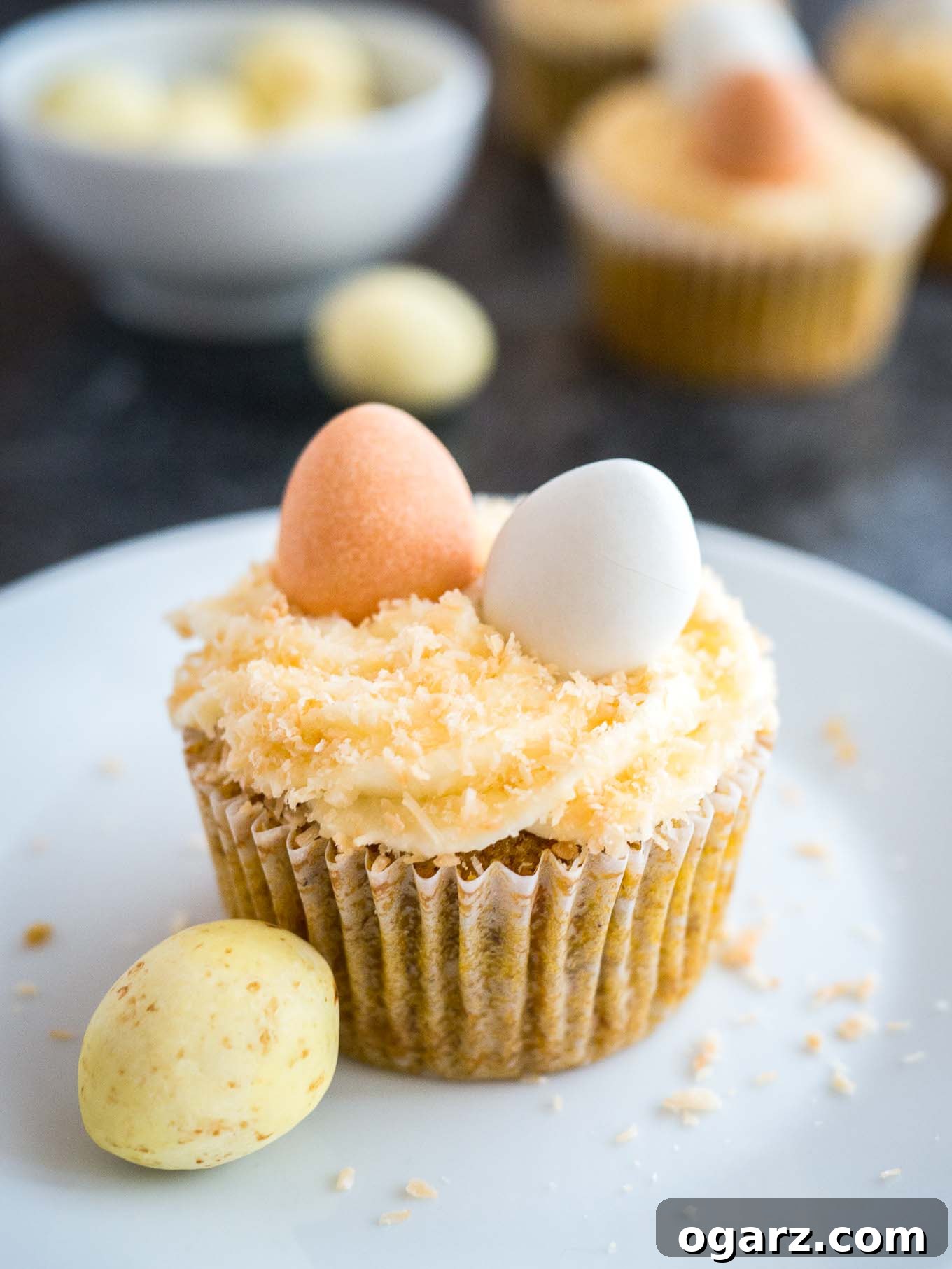 A delightful carrot cake cupcake, lavishly topped with cream cheese frosting, toasted coconut, and miniature chocolate eggs, presented on a white plate beside a decorative sugar egg.
