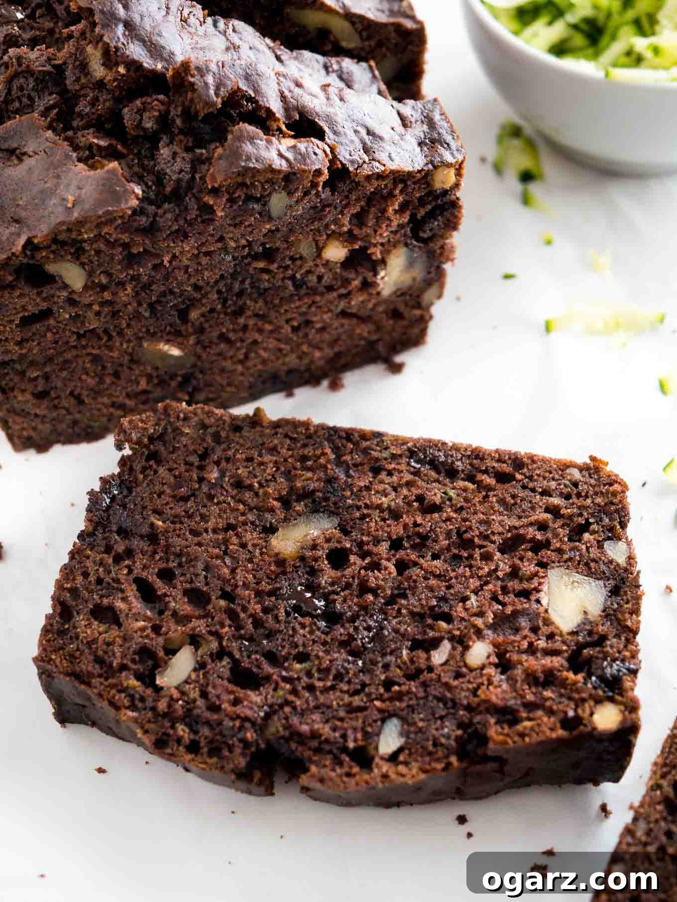 Decadent Chocolate Zucchini Bread 3 Close-up of a loaf of chocolate zucchini bread with two slices cut off, one standing one lying in front. There's a white bowl of shredded zucchini next to it.