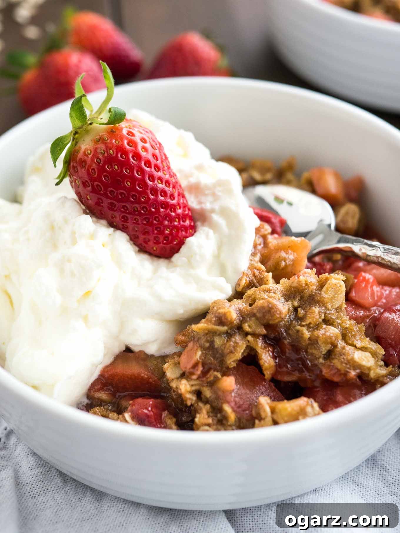 A beautifully presented Strawberry Rhubarb Crisp in a white bowl, generously topped with a dollop of fresh cream and a ripe strawberry, with a spoon ready for a taste.