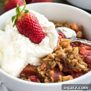 Strawberry rhubarb crisp in a white bowl topped with cream and a strawberry with a spoon in it.